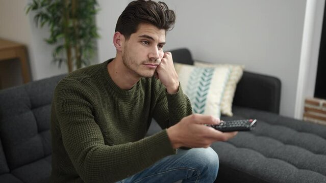 Young hispanic man watching tv sitting on sofa with boring expression at home