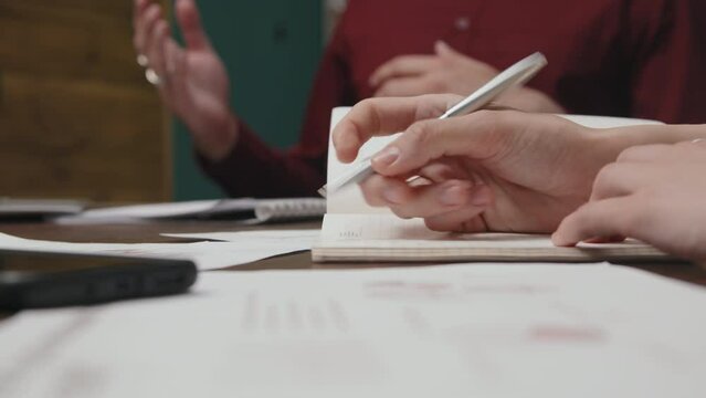 Close-up selective focus shot of hands of anonymous female assistant sitting in business meeting and rapidly writing in notebook, and unrecognizable person talking and gesticulating in background