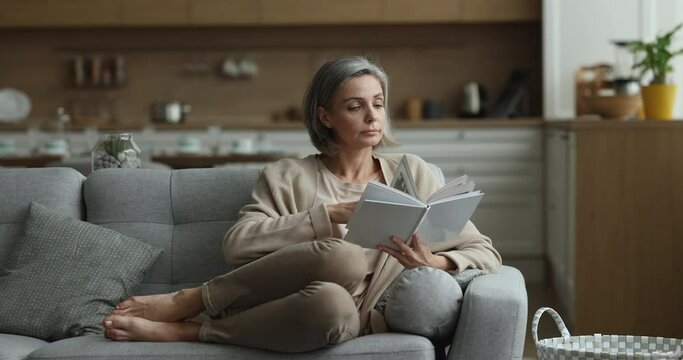 Focused middle aged woman relaxing at home, reading paper book, enjoying novel, leisure, studying new skills, getting knowledge, resting on comfortable couch at home