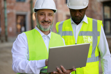 Portrait ofthe two builders together standing, looking at construction area with wireless laptop working on the project. Concept of business meeting and building process.