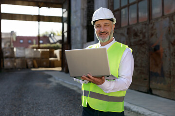 Portrait of the man builder in helmet and vest using wireless laptop for improving blueprints while standing at construction site. Concentrated bearded maleplanning system of building structures.