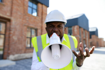 Portrait of man architect with loudspeaker standing at construction site. Angry african american foreman in protective helmet looking and aggressive shouting on camera.