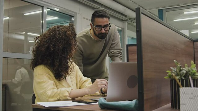 Medium shot of Caucasian female company worker sitting with laptop in cubicle in open-plan corporate office, and explaining something to Middle Eastern male colleague leaning on her desk