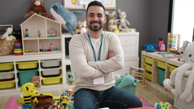 Young hispanic man teacher smiling confident sitting with arms crossed gesture at kindergarten