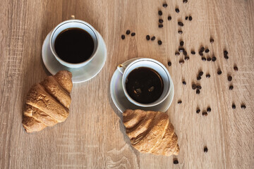Two cup of hot coffee in a plate and a croissants. Coffee beans are scattered on a wooden light...