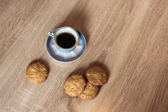A cup of hot fragrant black coffee in a saucer with cookies, on a wooden light brown textured table. Image for your creative design or illustrations.