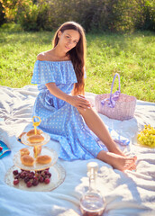 Picnic scene: sweet girl sitting on plaid in park. Outdoor portrait of adorable young woman in blue dress