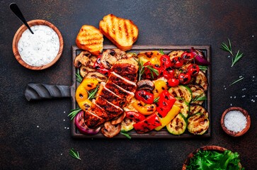 Grilled vegetables and chicken fillet. Paprika, zucchini, eggplant, mushrooms, tomatoes, onion and rosemary, served on rustic wooden cutting board, brown table background, top view