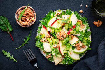 Waldorf salad with red and green apple, celery, lettuce, arugula and walnuts on plate, black table background, top view