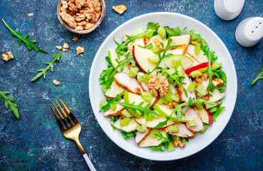 Waldorf salad with green and red apple, celery, lettuce, chicken fillet, arugula and crunchy walnuts on plate, blue table background, top view
