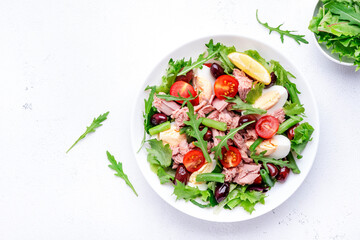 Nicoise salad with tuna, red tomatoes, boiled eggs, green beans and olives on plate, white table background, top view