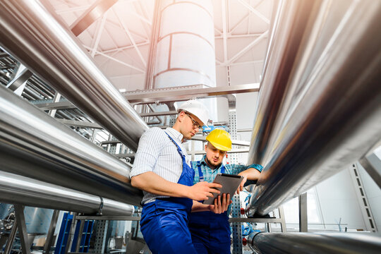 Two factory workers work with a tablet PC together. Stainless steel pipeline.