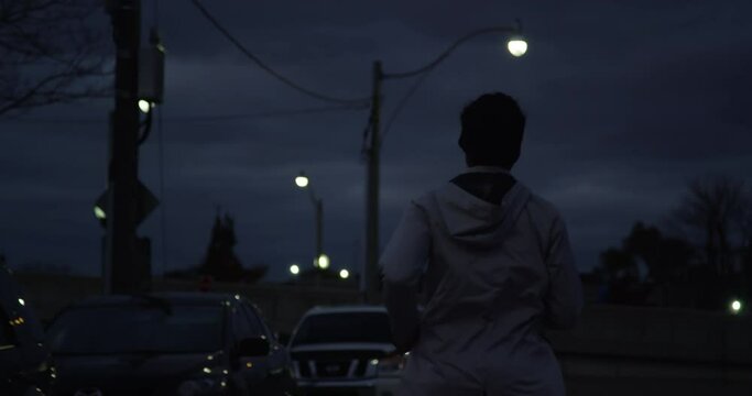Slow-motion of a female running at night down a dark street with city lights in the background