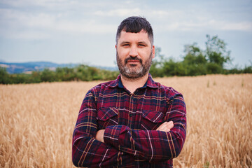 Naklejka na ściany i meble Arms crossed mature farmer man looking serious at camera standing at wheat field. Front view of folded agricultural rural worker with pensive attitude at harvest farm land. High quality photo