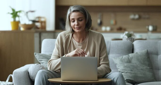 Positive middle aged 45 freelance woman using laptop computer, sitting on couch, working from home, typing, using online application, looking away, thinking on idea, getting serious, smiling
