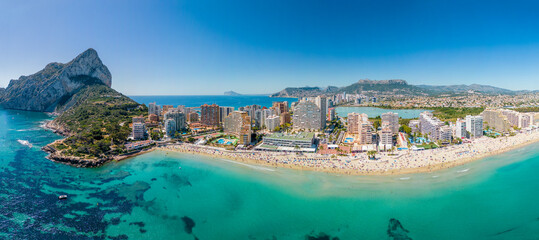 Wide Aerial Panorama of the Calpe beach with people on a clear summer day. Costa Blanca paradise riviera coastline with hotels and beaches with turquoise water.  El Peñón de Ifach, Calpe, Alicante. 