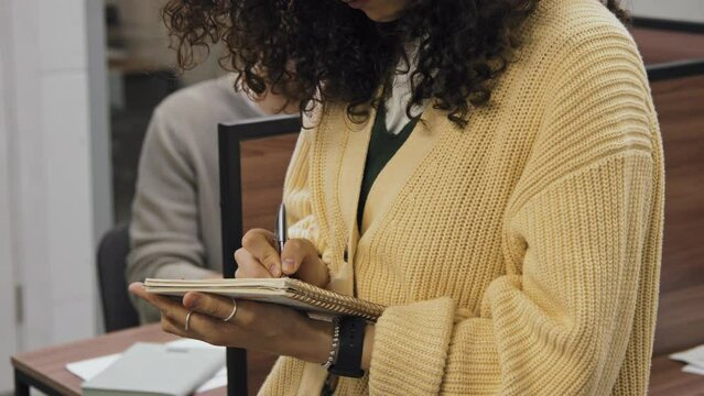 Close-up shot of hands of anonymous woman in yellow cardigan standing in middle of office, holding notebook and pen, and writing down ideas