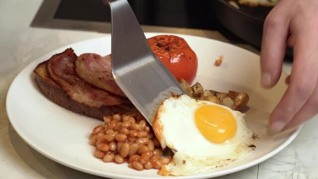 English breakfast on a plate. A cook puts a fried egg on a plate of a breakfast plate with beans, tomato bacon and toast.