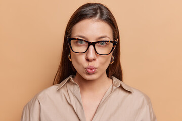Mysterious dark haired European woman looks attentively at camera keeps lips rounded wears spectacles and shirt poses against brown studio background. Lovely brunette female model stands indoors