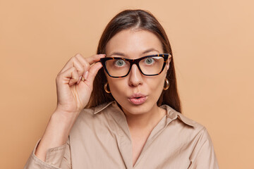Portrait of surprised woman stares impressed at camera keeps hand on rim of spectacles wears shirt...