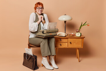 Happy redhead woman rejoices finishing working poses with typewrites on knees dressed formally poses in room with antique furniture and old items isolated over brown background. Retro office worker - obrazy, fototapety, plakaty