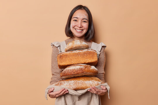Happy Asian woman holds stack of loaves of bread on napkin demonstrates yeast free organic food baked by herself smiles friendly has flour on face wears jumper and apron isolated over beige background