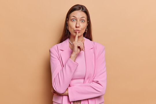Surprised brunette Euroepan woman makes silence gesture looks wondered at camera keeps quiet taboo sign wears formal pink jacket isolated over brown studio background. Body language concept.