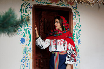 Fototapeta premium Charming smiling woman in traditional ukrainian handkerchief, necklace and embroidered dress standing at background of decorated hut. Ukraine, style, folk, ethnic culture