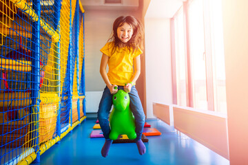 Little girl enjoys spending time in children's play center. Child jumps on bouncing inflatable rubber horse toy for kids. Good plaything for strengthening muscles, developing balance and fun.