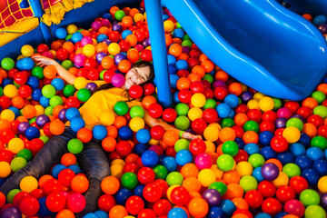 Little girl enjoys spending time in a dry pool with colorful plastic balls. The child lies on his...