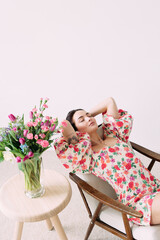 Young dreaming woman sitting in armchair with closed eyes next to vase with flowers bouquet on the table.