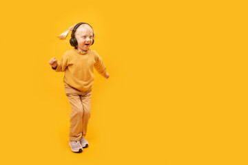 Full-length portrait of small blonde girl in yellow suit with headphones on yellow background. Child listens to music and dances.