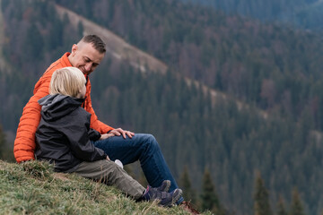 Naklejka na ściany i meble Father and son sit on hillside on the forest background. Weekend hike in mountains. Trekking and hiking with children.