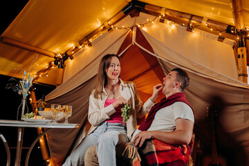Obraz na płótnie Canvas Happy lovely couple relaxing in glamping on summer evening and eating grapes near cozy bonfire. Luxury camping tent for outdoor recreation and recreation. Lifestyle concept