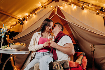 Obraz na płótnie Canvas Happy lovely couple relaxing in glamping on summer evening and drinking wine near cozy bonfire. Luxury camping tent for outdoor recreation and recreation. Lifestyle concept