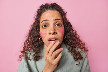 Fototapeta na wymiar Puzzled worried Brazilian woman with curly hair stares at camera keeps mouth opened applies collagen hydrogel patches under eyes to reduce wrinkles isolated over pink background. Beauty concept