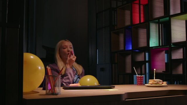 sad young female worker in the office celebrates a holiday with a sandwich cake with candles and a festive hat on her head
