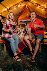 Fototapeta na wymiar Happy lovely couple with Welsh Corgi Pembroke dog relaxing in glamping on evening and drinking wine near cozy bonfire. Luxury camping tent for outdoor recreation and recreation. Lifestyle concept