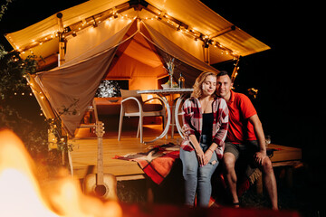 Obraz na płótnie Canvas Happy lovely couple relaxing in glamping on evening near cozy bonfire. Luxury camping tent for outdoor recreation and recreation. Lifestyle concept