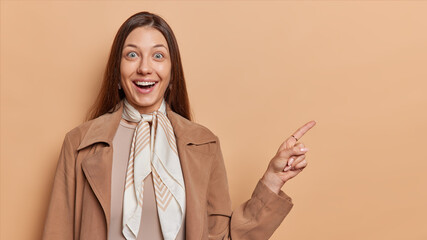 Cheerful surprised young woman with dark straight hair points index finger at upper left corner...