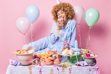 Hungry positive woman dressed in pajama bites delicious doughnut poses near messy festive table rests after party surrounded by inflated balloons isolated over pink background. Birthday celebration