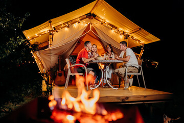 Happy group of friends relaxing in glamping and drinking wine on summer evening near cozy bonfire. Luxury camping tent for outdoor recreation and recreation. Lifestyle concept