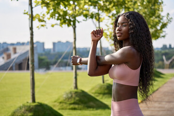 Young adult confident beautiful fit sporty slim active black African ethnic woman runner model wearing sportswear top arm muscles warming up exercise training standing in city park outdoors.
