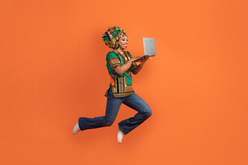 Happy black woman in african costume jumping with open laptop