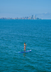 SUP Stand up paddle board. Blond girl sailing on paddle board in blue sea. Woman on summer holidays vacation lifestyle. Batumi city on background. Georgia country.
