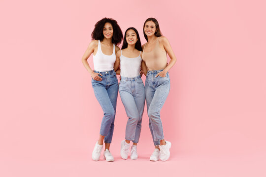 Happy three multiracial girls embracing and smiling to camera, posing in studio over pink background, full body lenght