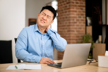 Tired stressed asian businessman having neck pain, touching and massaging neck, sitting at workplace with laptop at home