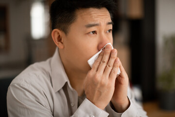 Influenza. Sick middle aged asian man blowing runny nose sneezing, suffering from cold and high fever, feeling unwell