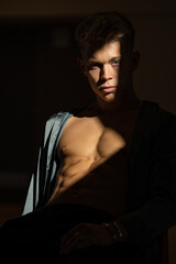 Fototapeta premium A muscular man in a gray satin shirt sitting on a dark background with falling sunlight