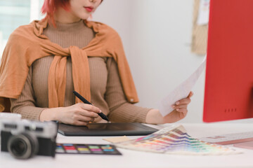 A creative and stylish young female freelance designer is using a tablet and stylus pen while...